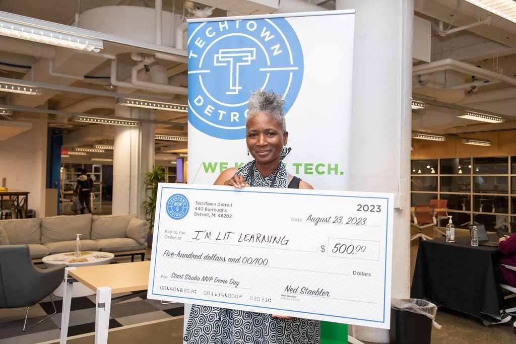 A woman holds up a large check from TechTown Detroit for $500 for her startup, I'M LIT Learning