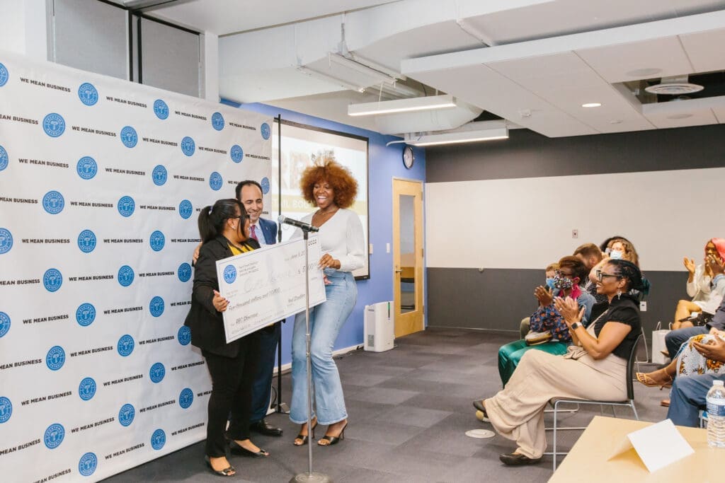 A woman smiles as she receives a large check for $5,000 for her business, Cure Nailhouse. One woman and one man are handing off the check and smiling. The three of them are standing before a seated group of people, who are smiling and clapping.