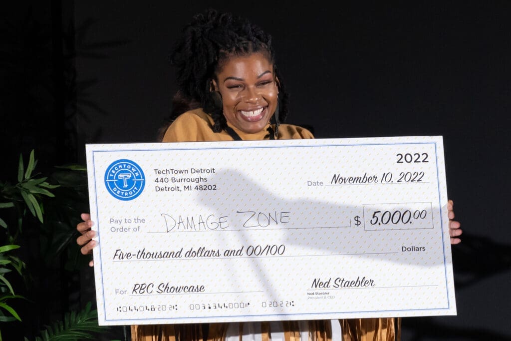 A woman smiles and holds a check for $5,000 from TechTown Detroit for her Detroit-based rage room and axe-throwing business, The Damage Zone.
