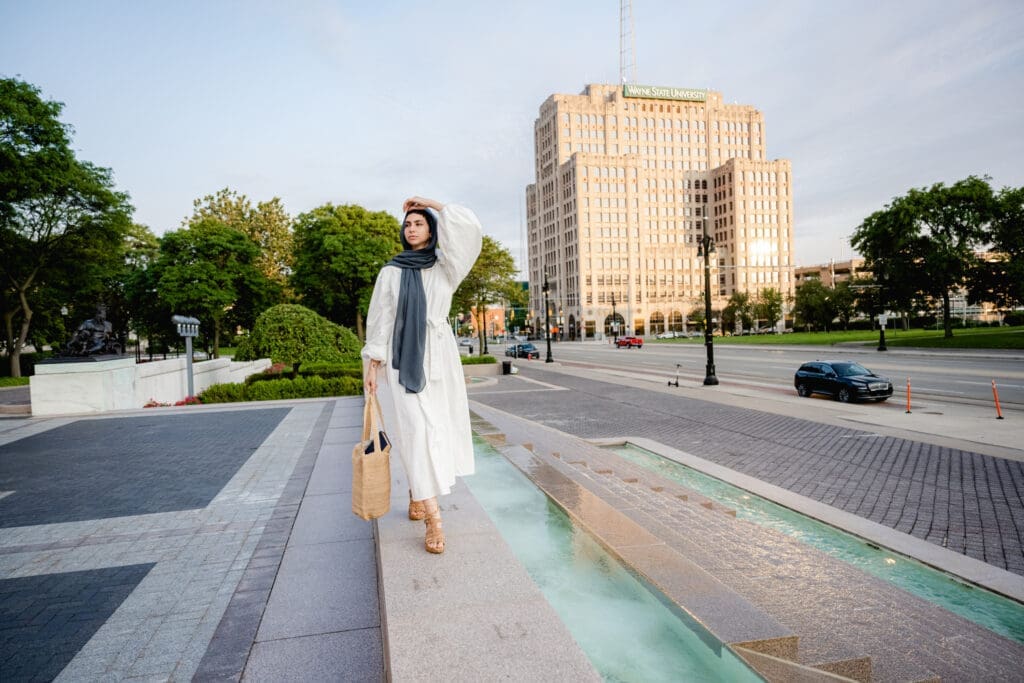 A woman poses outside, wearing a dark grey hijab, a long-sleeved white maxi dress and tan strappy sandals. She holds a tan tote bag.