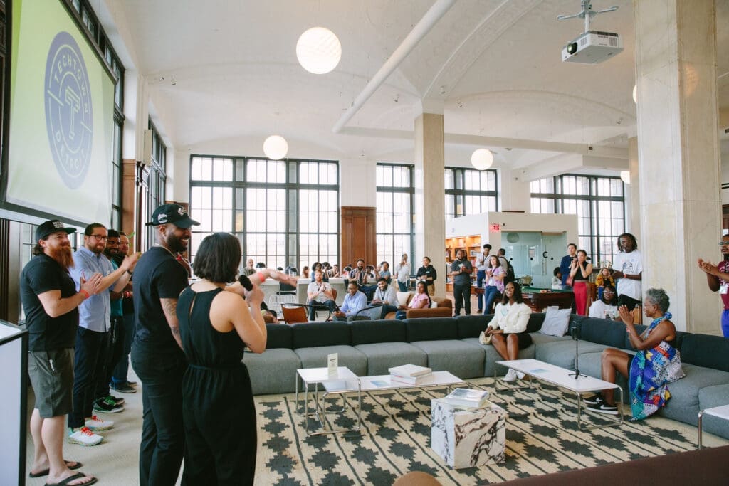 A man smiles next to a woman holding microphone who is speaking to a crowd of people standing and sitting on couches and chairs in a well-lit event space. Behind the man stands four men standing under a projector hanging from the ceiling. The projector shows TechTown Detroit's blue and white logo.