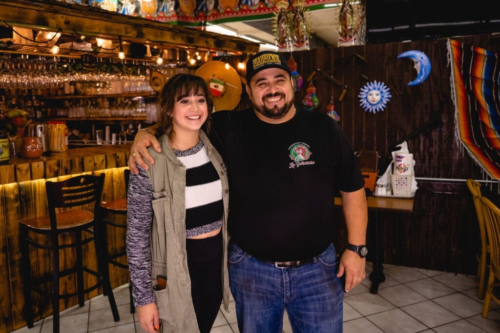 A woman and man smile for their photograph. The man has his arm around the woman's shoulder.