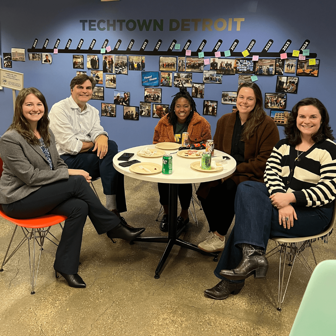 Five people sit around a a table, smiling for a photo. There are plates of food and beverages on the table.