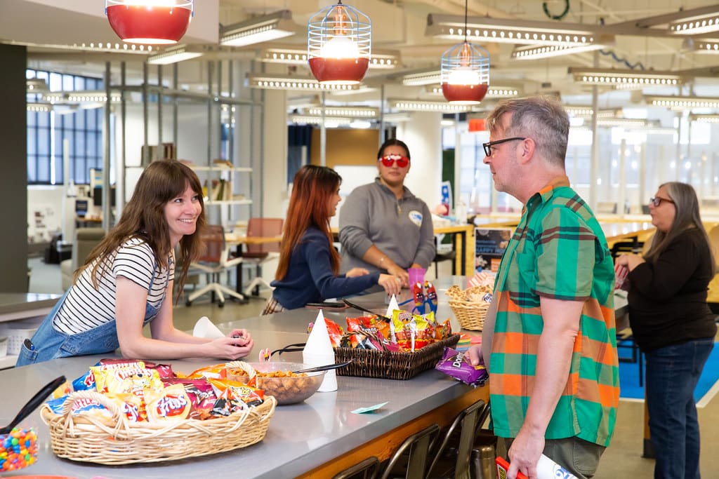 A person leans over a table full of snacks inside a coworking office and converses with another person standing in front of the table.